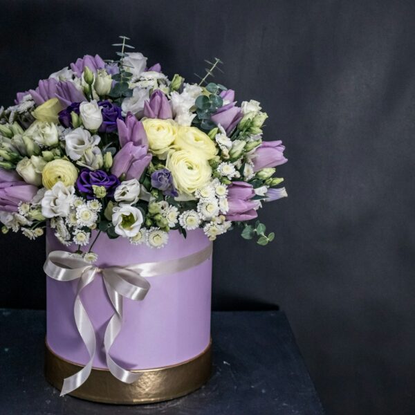 White and purple flower bouquet in a lavender box with a white ribbon and a dark grey background